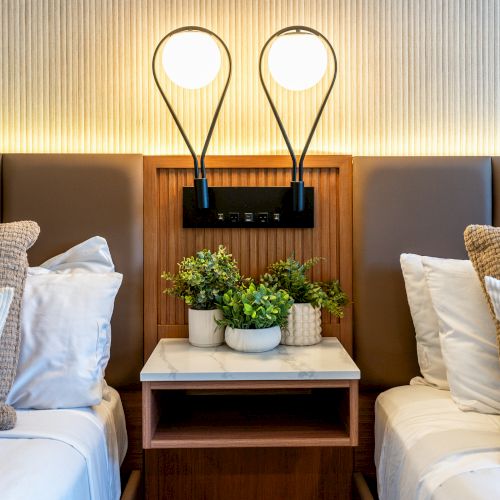 A modern bedroom features two beds with striped pillows, a shared nightstand, decorative plants, and two wall-mounted lights above.