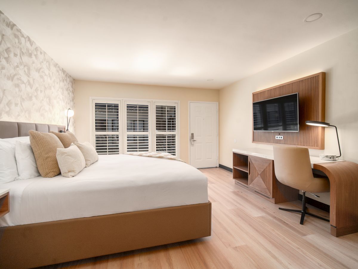 A modern hotel room features a large bed, bedside tables, a flat-screen TV on a wall-mounted desk, a chair, and a window with shutters.