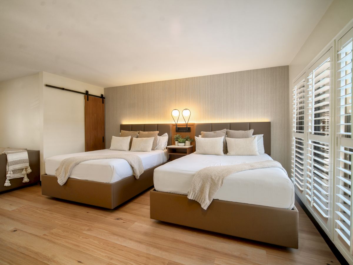 A bedroom with two beds, a chair, wooden flooring, a sliding barn door, and ample natural light from the large window with white shutters.