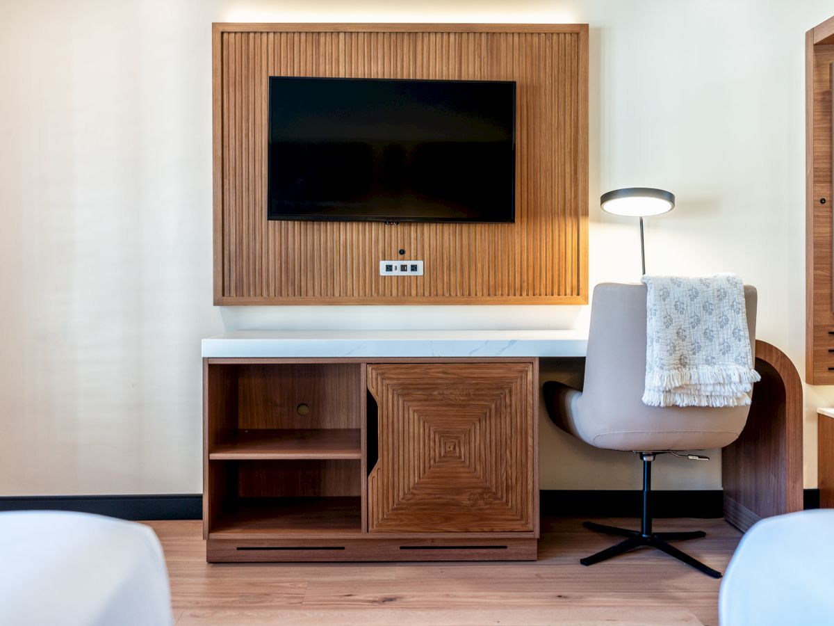 A hotel room features a TV on a wooden panel, a desk with a lamp, a chair, and a blanket draped over the chair.