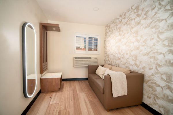 A cozy, small room with a couch, a wall mirror, an air conditioner, and patterned wallpaper. The space appears well-lit and minimally decorated.