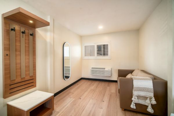 A small room with a brown couch, white blanket, wall mirror, coat rack, bench, wooden floor, window with shutters, and wall-mounted air conditioner.