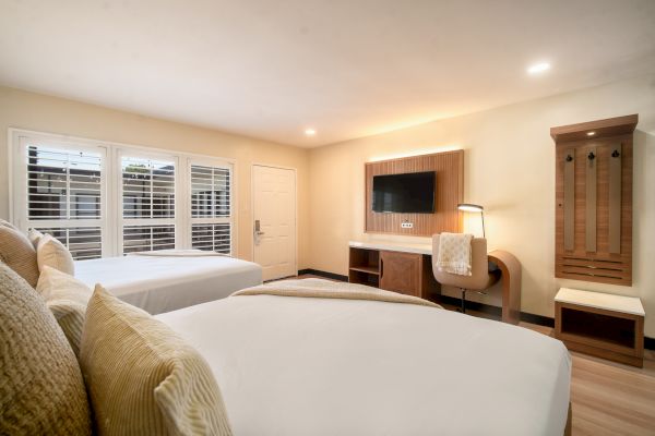 A hotel room with two beds, a wall-mounted TV, a desk, a chair, and a wooden wardrobe. The room has a clean, modern design with soft lighting.