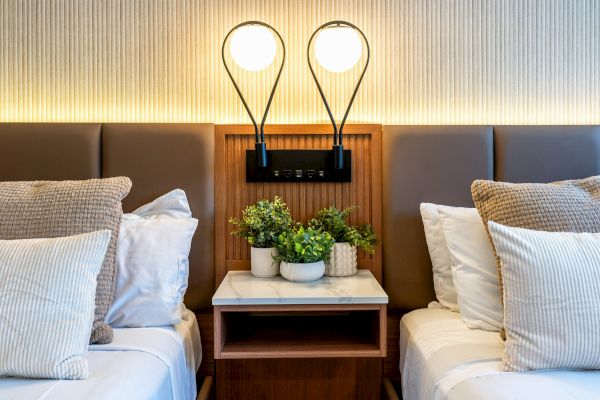 A hotel room features two beds with a shared nightstand holding potted plants. Two modern lamps are mounted above the nightstand, illuminating the area.