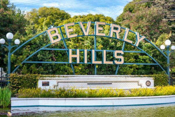 The image shows the iconic Beverly Hills sign surrounded by greenery and lamps, with a small body of water in front of it.