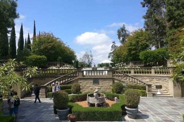 The image shows an outdoor garden with stone staircases, well-manicured greenery, and a central fountain under a partly cloudy sky.