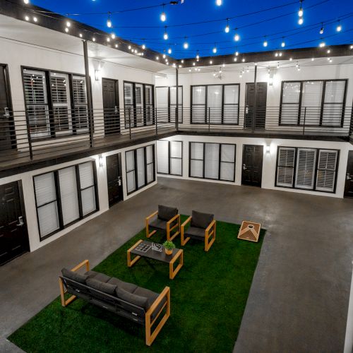 A modern courtyard with string lights, patio furniture on a green turf, surrounded by black and white building walls with multiple doors and windows.