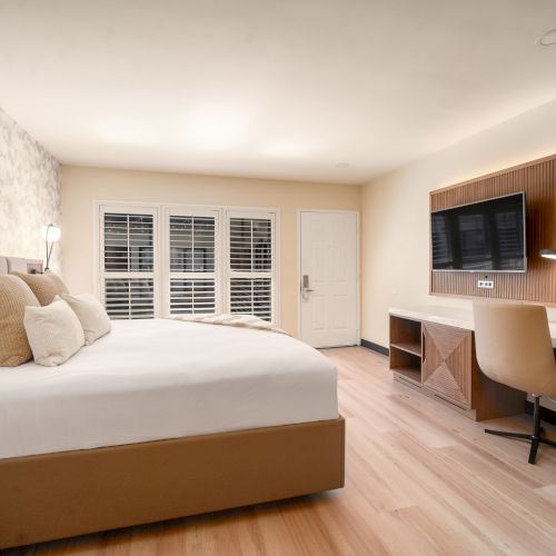 A modern hotel room features a large bed with pillows, a desk with a chair, a television, and large windows with shutters, plus minimalist decor and lighting.