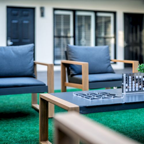 Outdoor seating area with two chairs, a table, a small plant, and a board game on artificial grass in a modern courtyard is seen.