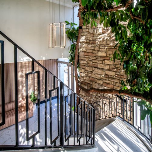 A staircase with a sleek metal railing leads past a large indoor plant and a stone accent wall, illuminated by natural light and modern hanging lights.