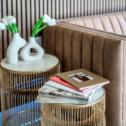 The image shows a stylish living room corner with a beige leather couch, two round marble-top tables, white tulip vases, and a stack of books and magazines.