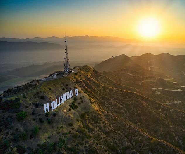 An aerial view of the Hollywood Sign in Los Angeles at sunset, perched on a hillside with lush greenery and a clear sky in the background.